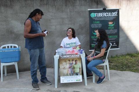 Duas mulheres estão sentadas próximo a uma mesa onde há cartaz e banner com informações sobre tuberculose. Um homem está ao lado, em pé. Eles conversam. #Pracegover