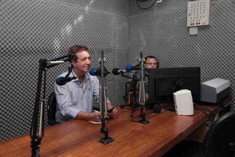 O estúdio da rádio com microfones, monitor de computador e duas pessoas falando. #Pracegover