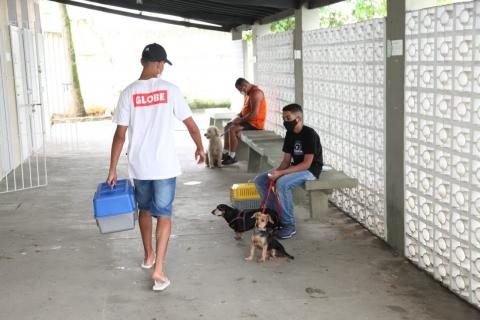 Corredor com bancos onde estão sentados dois homens segurando seus cachorros. Um homem está de costas, saindo, com uma caixa própria para pet. #Paratodosverem