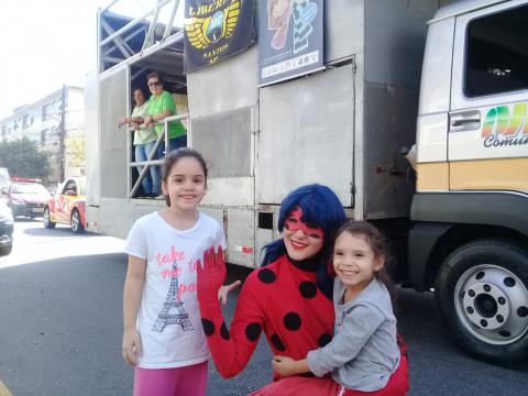 Mulher vestida de personagem infantil posa para foto com duas crianças á frente de caminhão