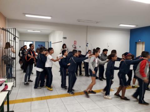 Crianças simulam saída da escola em fila e com as mãos umas nos ombros das outras. #Pracegover