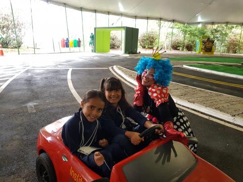 estudantes estão em mini carro com mulher vestida de palhaça ao lado #pracegover 