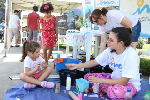 Mulher e menina sentadas sobre tapete na praça manuseiam materiais como corda, cola e tinta