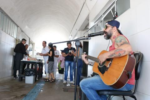 Homem toca violão e canta ao microfone sentado em cadeira, enquanto púvluco se serve de ceviche