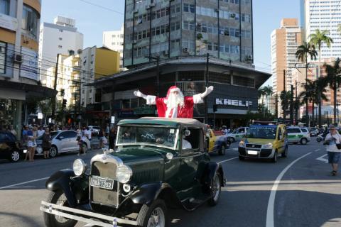 #pracegover Em pé dentro de calhambeque, Papai Noel acena de braços abertos à frente da Praça Independência