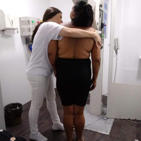 Mulher está fazendo exame em mamógrafo. Uma auxiliar, vestida de branca, a auxilia. #Pracegover