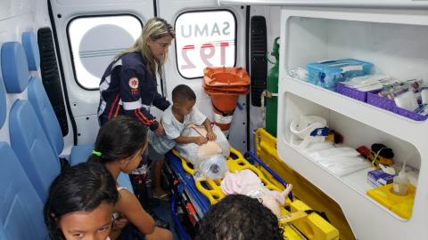 #pracegover Dentro de ambulância, enfermeira uniformizada observa menino fazendo masssagem cardíaca em boneco