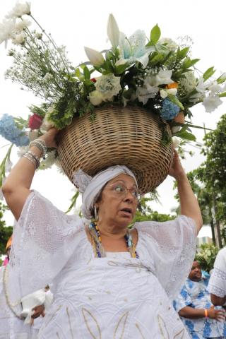 #pracegover Mulher de branco carrrega na cabeça um vaso com flores brancas