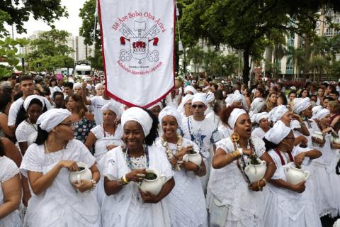 #pracegover Dezenas de mulheres de btanco com potes brancos na mão caminham pela orla da praia