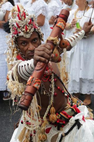 #pracegover Homem travestido de orixá, com ornamentoa africanos, posa sério para foto com instrumento cilíndrico na mão