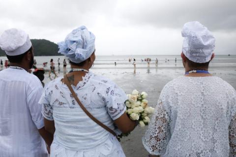 #pracegover Dois homens e uma mulher com flores na mão aparecem de costas olhando em direção ao mar