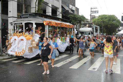 #pracegover Sobre a rua molhada, bonde circula com centenas de pessoas atrás