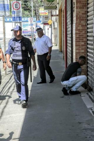 Homem fecha porta de ferro de estabelecimento em rua que já tem outras casas fechadas. Guardas municipais estão ao lado. #Paratodosverem