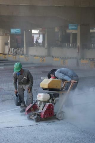 HOmens uniformizados, com equipamentos de proteção como capacetes e luvas, quebram o piso antigo. Um deles opera uma máquina. #Paratodosverem