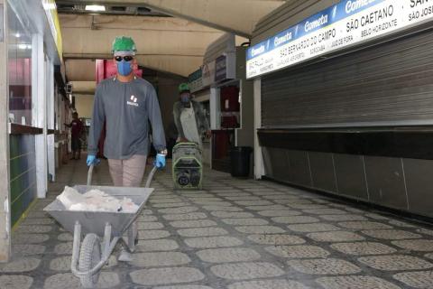 Dois operários caminham em um corredor. O da frente empurra um carrinho de mão com entulho. O de trás empurra uma máquina. Eles estão bastante distantes um do outro. #Paratodosverem