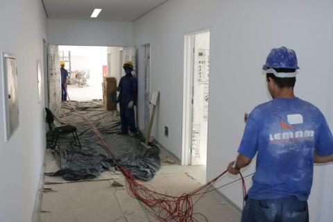 Dois operários estão trabalhando em corredor de futuro hospital. Eles seguram diversos fios. #Paratodosverem