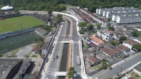 Vista aérea da avenida renovada, com um campo de futebol à esquerda e prédios e casas à direita. #Paratodosverem