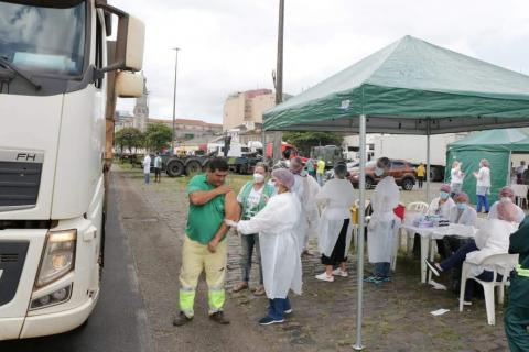 Caminhoneiro está na calçada sendo vacinada por uma pessoa com avental branco, touca, máscara e luva. Ao lado deles, à esquerda, está o caminhão. Do lado direito, uma tenda onde se concentram os serviços de imunização. #Paratodosverem