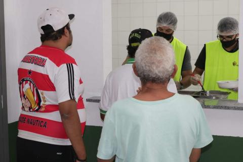 #pracegover Três homens formam fila para receber comiga servida por dois homens uniformizados em balcão