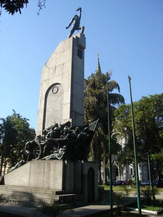 No centro uma praça com um monumento cercado por árvores - #paratodosverem