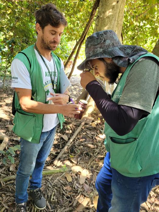 dois agentes uniformizados estão em área rural observando larvas em recipiente. #paratodosverem