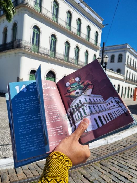 mão segura livro em primeiro plano com página aberta no Museu Pele. Imóvel real está ao fundo. #paratodosverem