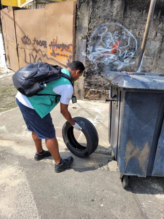agente uniformizado e usando luvas carrega pneu velho que acumula água. #paratodosverem