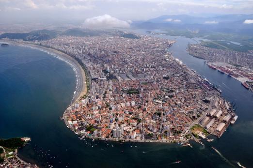 silhueta da ilha de São Vicente, vista do alto, com a ponta da praia em primeiro plano, a orla de Santos do lado esquerdo, e o porto, do direito. #Paratodosverem