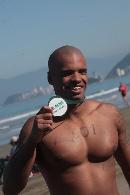 Lucas Barbosa está de costas para o mar e segura medalha de participação sorrindo para a foto. #paratodosverem