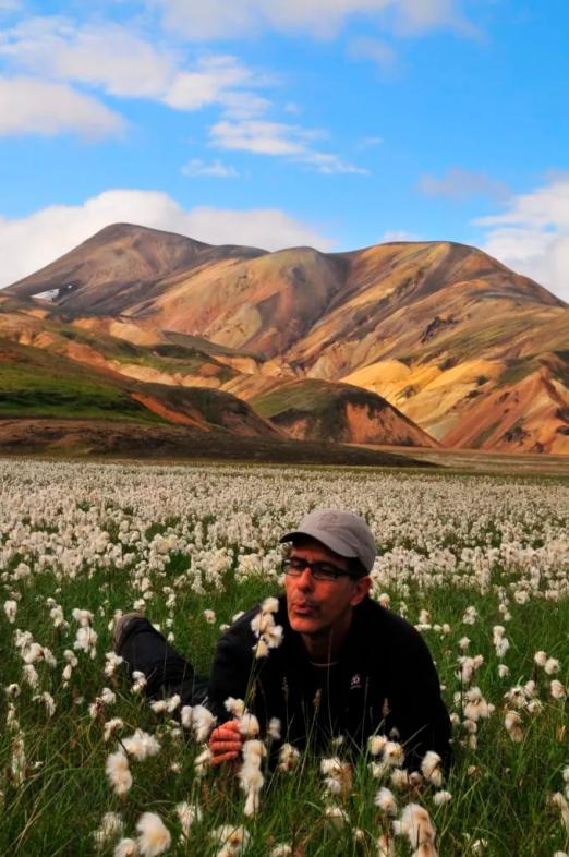 jornalista está no meio de um campo de flores com uma câmera fotográfica na mão. Ao fundo, um morro. #paratodosverem