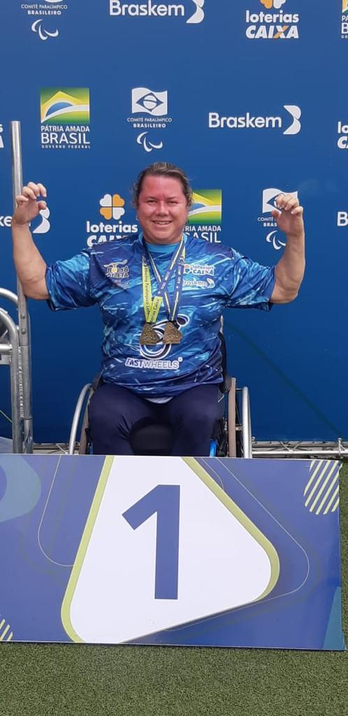 Atleta ergue braços com medalhas em pódio de primeiro lugar #paratodosverem