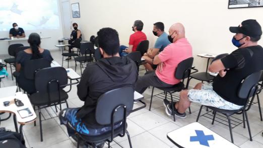 Agentes sentados em cadeiras observam instrutor com telão ao fundo. #pratodosverem