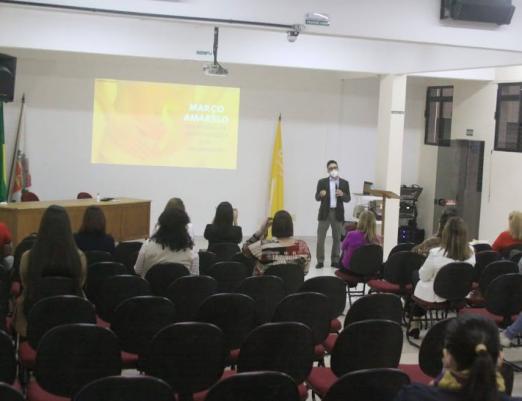 Várias pessoas sentadas assistem homem falando com telão ao fundo. #paratodosverem