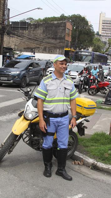 Ricardo está na rua, próximo à via e a frente da moto que usa para trabalhar. Há carros parados no semáforo ao fundo. #paratodosverem