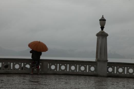 Pessoa está passada atrás de mureta na Ponta da Praia em um dia chuvoso. Ela segura um guarda-chuva aberto. #Paratodosverem