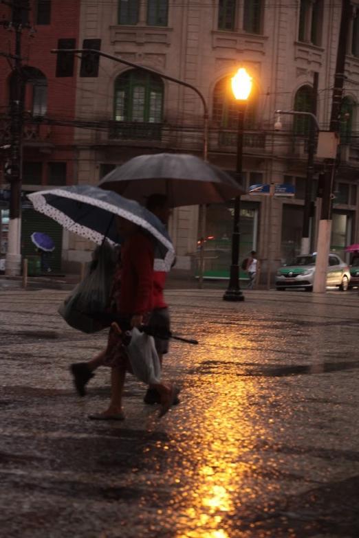 Duas pessoas caminham com guarda chuva em rua à noite. #Paratodosverem