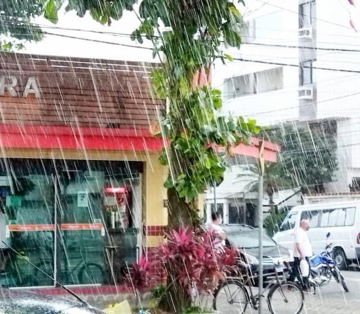 chuva caindo em rua. com estabelecimento comercial ao fundo e pessoas na rua. #paratodosverem