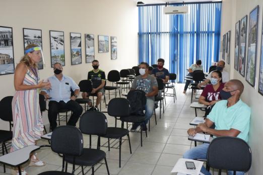 Sala de aula com pessoas sentadas em cadeiras espaçadas entre si. Mulher que está em pé fala ao grupo. #Paratodosverem