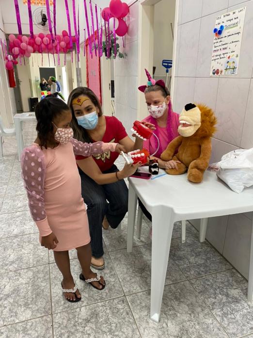 criança usando máscara mexe em bicho de pelúcia que está sobre uma mesa. O ambiente está decorado com balões de gás cor-de-rosa. Ao lado da menina há duas mulheres, uma deles usa uma fantasia infantil. #paratodosverem