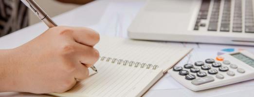 close de mão segurando caneta e escrevendo em caderno. Há uma calculadora e um notebook sobre a mesa. #paratodosverem