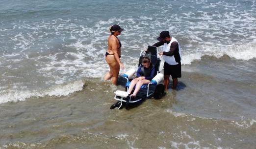 jovem está sentada em cadeira anfíbia tomando banho de mar. Técnico controla a cadeira. A mãe da jovem está em pé ao lado. Eles estão no mar. #paratodosverem 