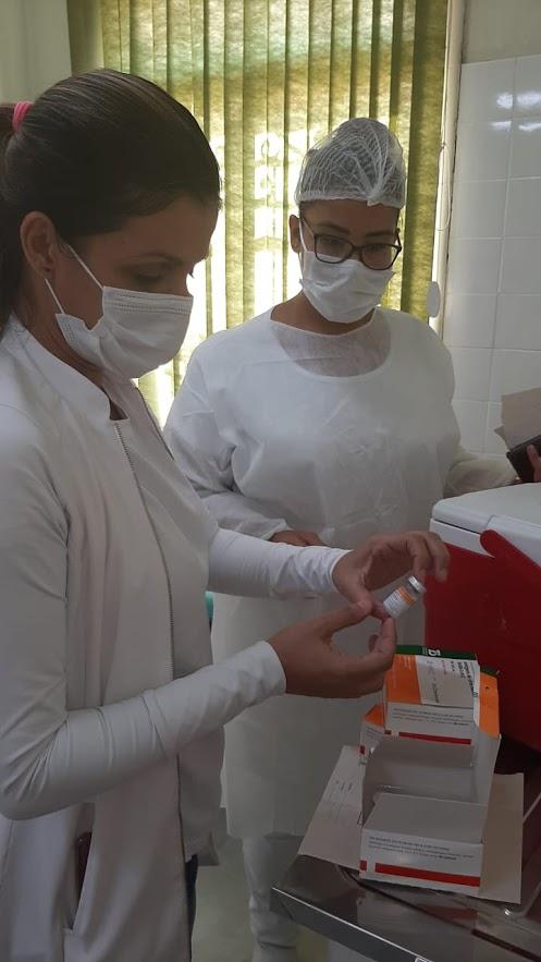 Duas mulheres vestidas de branco e usando máscara e luva mexem em caixa com doses de vacina. #Paratodosverem