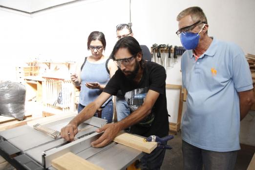 três pessoas durante curso de marcenaria olham para professor operando instrumento pra cortar madeira #pracegover 