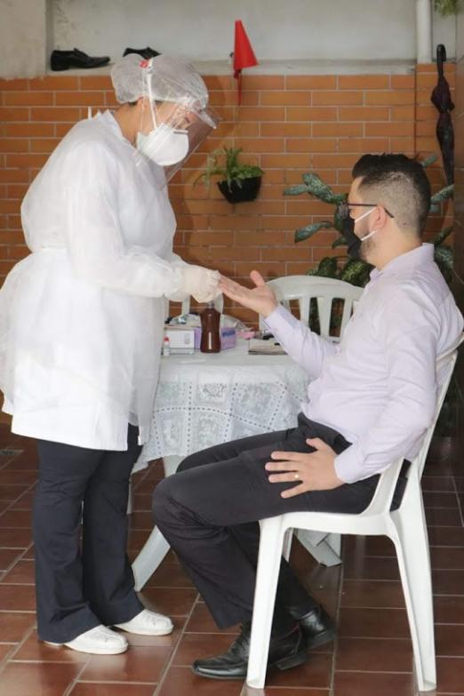 Mulher paramentada com avental, luva, gorro e máscaras faz teste em um homem sentado. #Paratodosverem