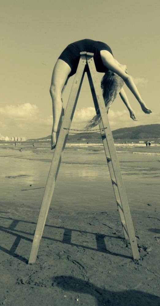 Uma escada está aberta na areia da praia. Uma mulher está com seu dorso flexionado sobre o topo da escada, de modo que seus braços estão de um lado da escada e suas pernas de outro. #Paratodosverem