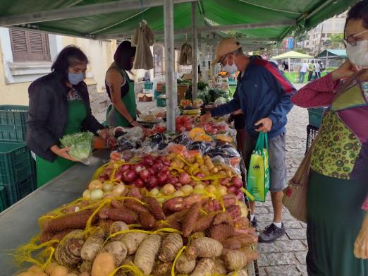 banca em feira com pacotes de batatas doces, cebolas e outros itens. Pessoas circulam em torno.  #paratodosverem