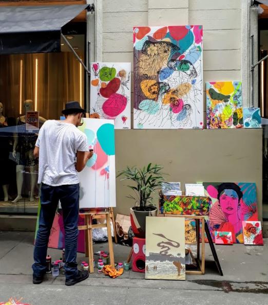 Artista pinta quadro em cavalete armado na rua. Ele está de costas. Há vários quadros encostados na parede ao fundo. #Paratodosverem