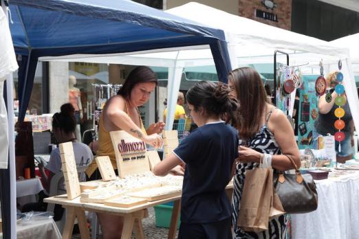 Duas mulheres olham produtos em banca de feira de artesanato. #Paratodosverem