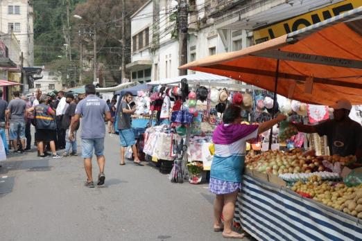 Feira está armada em rua. Pessoas estão andando na rua ou paradas em frente a barracas. #Paratodosverem