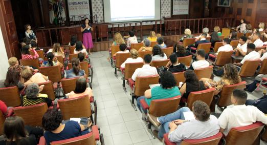 Auditório com poltronas lotado. Ao fundo, uma mulher fala ao público. #Pracegover